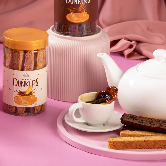 Almond House Almond Dunkers jars placed beside a white teapot, a cup of tea, and assorted almond biscotti on a pink background.
