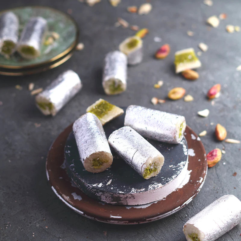 A plate of almond and pistachio rolls coated in edible silver leaf, with a few pieces scattered around, on a dark surface. Some of the rolls are cut open to reveal the pistachio filling inside.