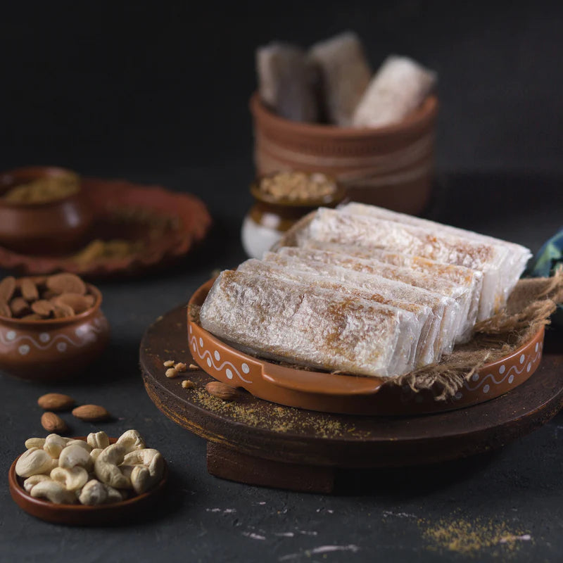 Traditional Indian sweet Bellam Pootharekulu neatly arranged on a rustic clay plate, surrounded by cashews and almonds in earthen pots.