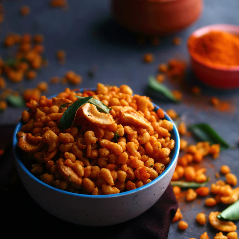 A bowl of Almond House Boondi snack garnished with curry leaves and cashews, surrounded by scattered boondi and spices.