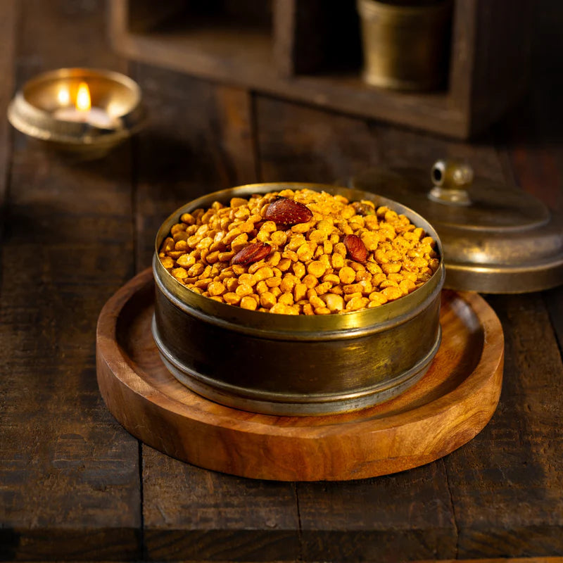 Almond House Chabina, a crunchy chickpea snack with almonds, served in a vintage metal container on a wooden platter, with a lit oil lamp in the background.