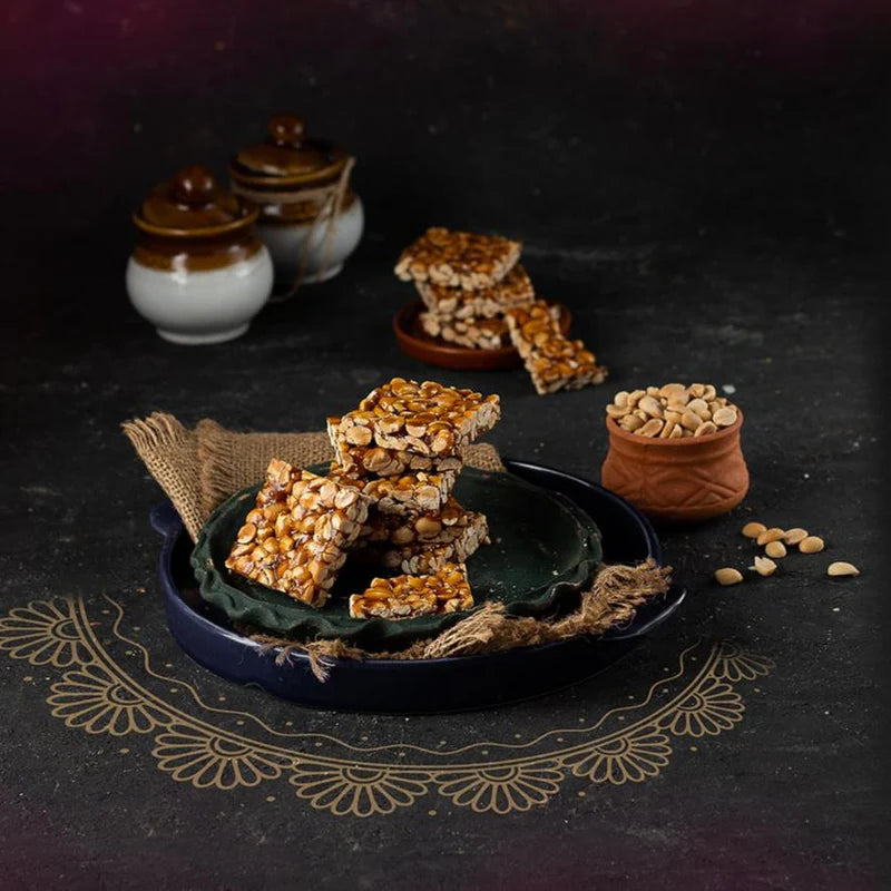 Almond House Groundnut Chikki made with roasted peanuts and jaggery, served on a rustic dark background with traditional Indian decor elements.