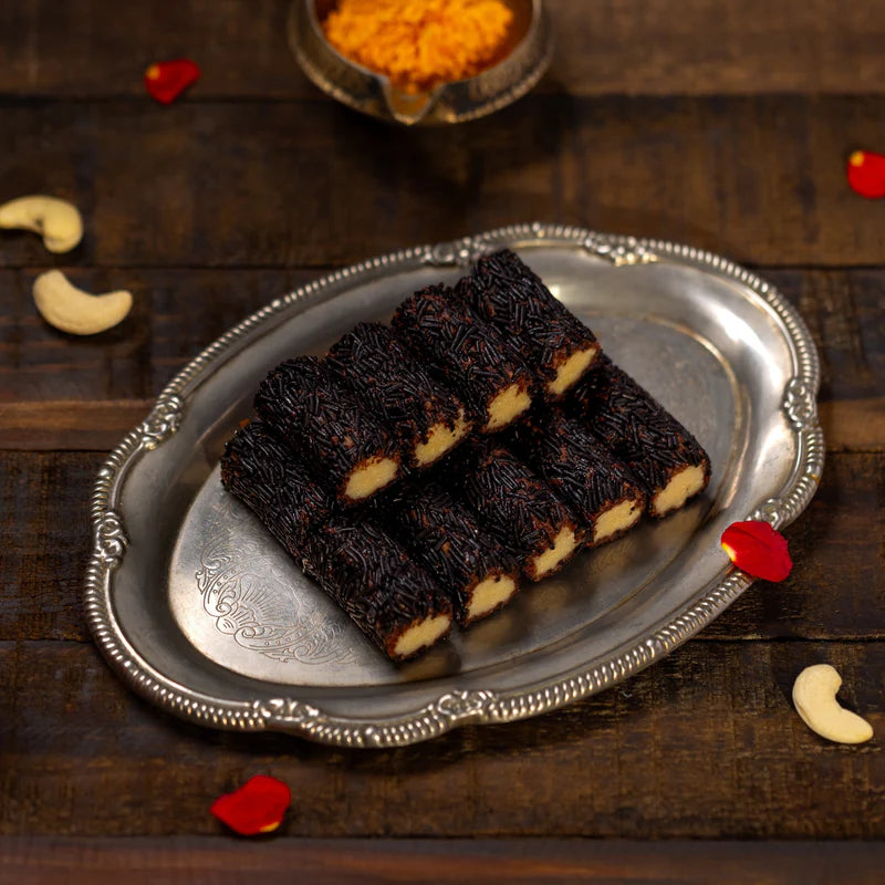 Almond House Kaju Flakes Roll with a creamy cashew filling, coated in chocolate sprinkles and presented on an ornate silver tray with rose petals and cashews.