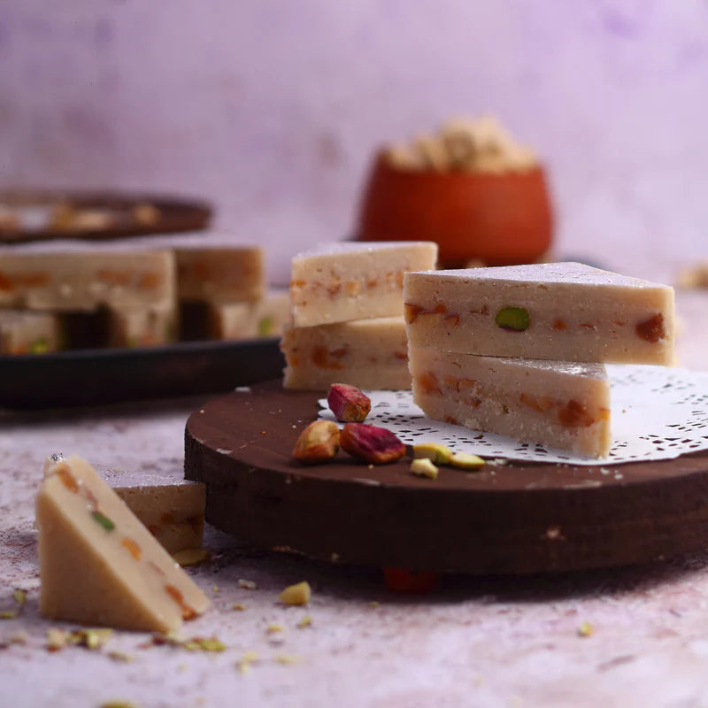 Triangular slices of Kaju Pista Sandwich stacked on a wooden tray, featuring rich dry fruit filling and a silver leaf topping.