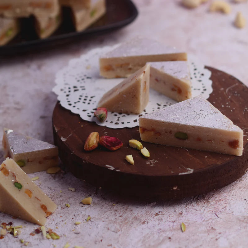 Close-up of Kaju Pista Sandwich, a traditional Indian sweet with cashew base and pistachio filling, served on a wooden platter with decorative paper.