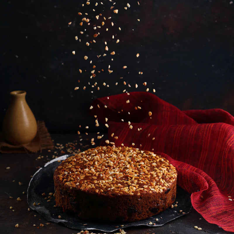 Almond House Plum Cake being sprinkled with chopped nuts, styled with a red cloth and dark background.