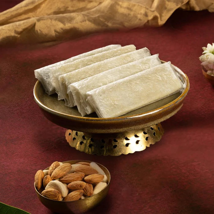 A golden stand showcasing Almond House Sugar Pootharekulu, traditional Indian sweet rolls, on a red surface, accompanied by a bowl of almonds, jasmine flowers, and draped beige fabric in the background.