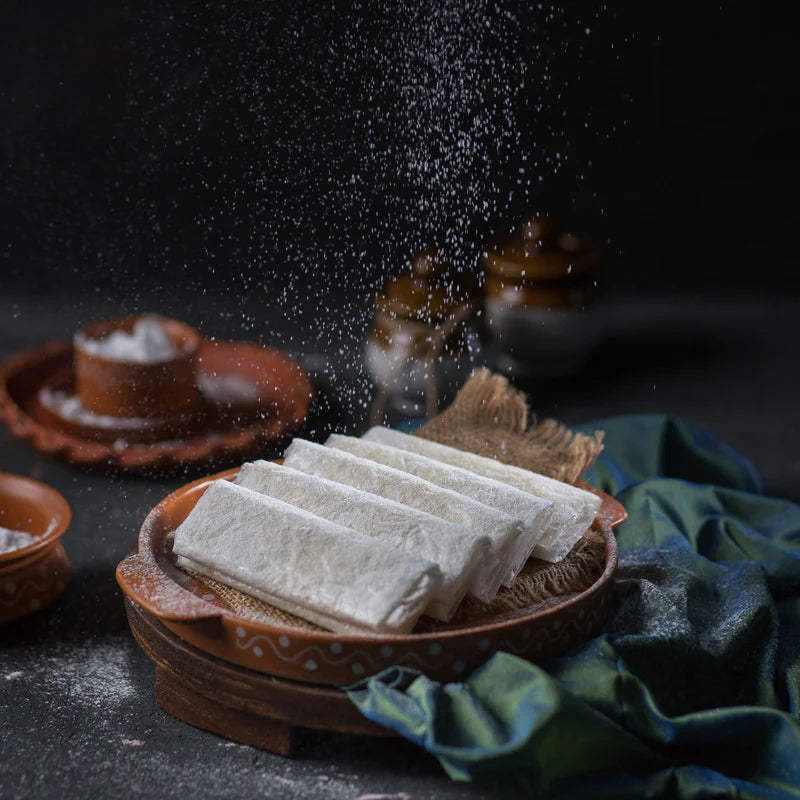A rustic clay plate of Almond House Sugar Pootharekulu, traditional Indian sweet rolls, being dusted with powdered sugar, surrounded by clay pots and a green-blue cloth, with burlap fabric in the background.