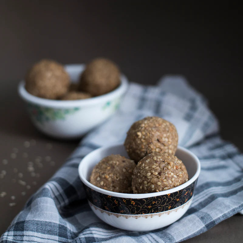Two white bowls filled with Almond House Till Laddu round sesame seed sweet balls are placed on a gray plaid cloth, with one bowl in the foreground and the other in the background.
