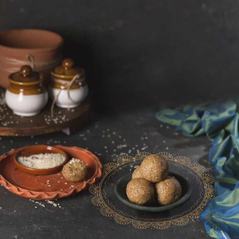 A plate with Almond House Till Laddu is placed next to a smaller dish containing sesame seeds and another ladoo, with jars and a cloth in the background.