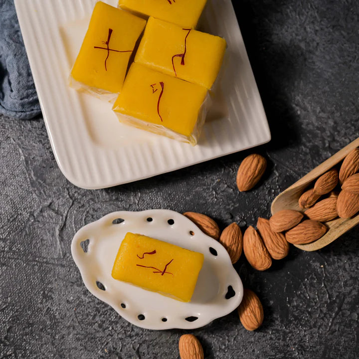 Vellanki Foods Badam Halwa sweets arranged on a plate, with a piece on a dish beside scattered almonds against a dark textured background.