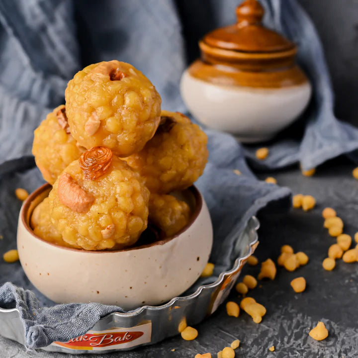 A bowl stacked with golden, Boondi Laddu sweets topped with nuts, surrounded by scattered ingredients on a textured background.