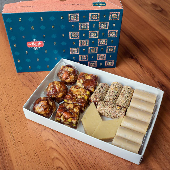 A variety of Indian sweets in a white box, alongside a decorative blue box, showcasing treats like bars, rolls, and nutty confections.