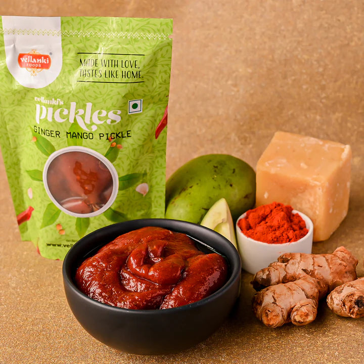 Vellanki Foods Ginger Mango Pickle in a black bowl with raw mango, ginger roots, red chilli powder, and jaggery, displayed in front of branded packaging.
