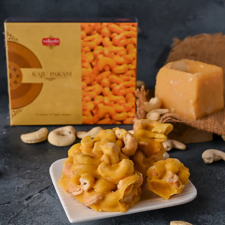 A plate of cashew brittle, called Vellanki Foods Kaju Sweet (Kaju Pakam), sits in front of its box, with cashews and a block of jaggery arranged on a dark surface in the background.