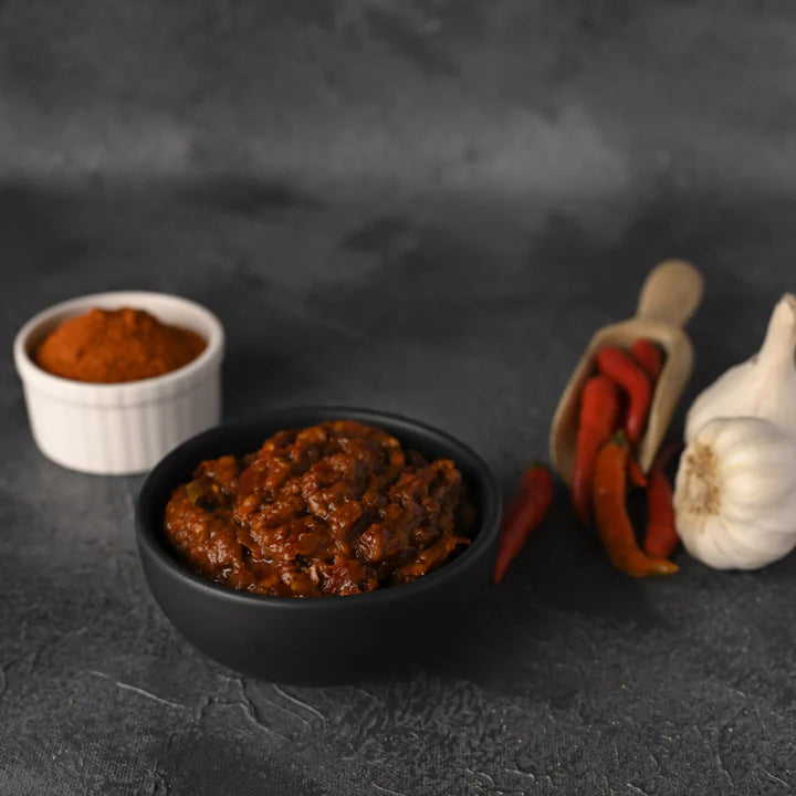 Vellanki Foods Red Chilli Pickle in a black bowl with garlic, red chillies, and spice powder.