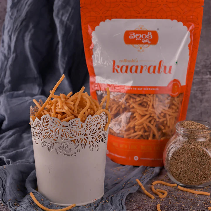 Vellanki Foods Vampoosa (Kaaralu) in a decorative white container with a branded snack pack and ajwain seeds displayed in the background on a fabric surface.