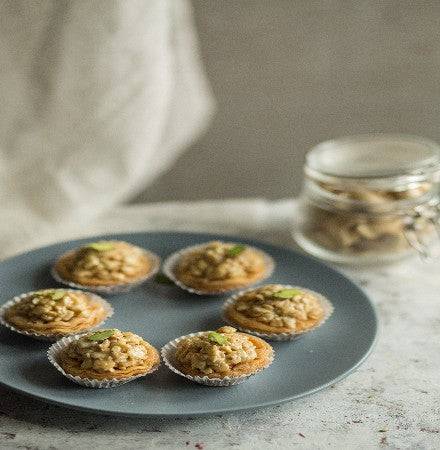 Side view of Almond House Honey Cashew Tarts on a blue plate, garnished with crushed cashews and pistachios, with a jar of cashews in the background.