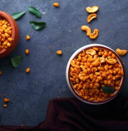 A bowl of Almond House Boondi snack garnished with curry leaves and cashews, surrounded by scattered boondi and spices.