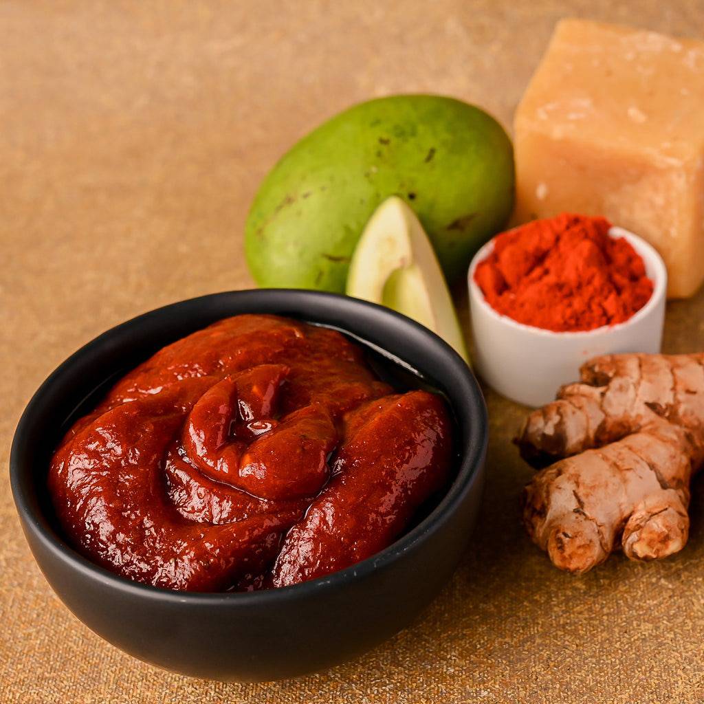 Vellanki Foods Ginger Mango Pickle in a black bowl with raw mango, ginger roots, red chilli powder, and jaggery.