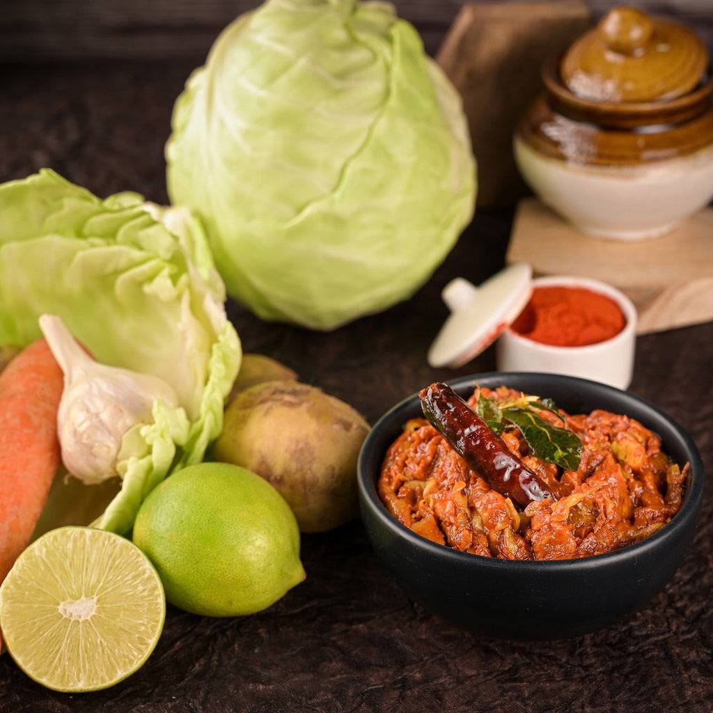 A bowl of spicy Vellanki Foods Mixed Veg Pickle and fresh vegetables like lime, cabbage, garlic, and carrot arranged around it.